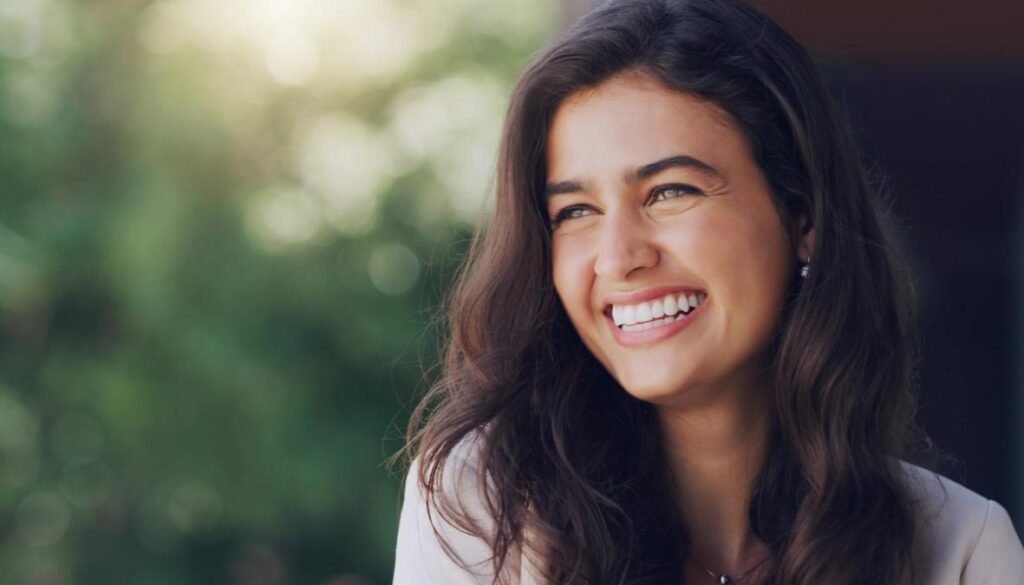 A joyful woman with long dark wavy hair smiles while looking to the side.