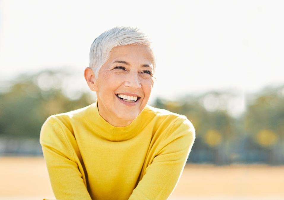 Smiling mature woman with short white hair in a yellow sweater outdoors.