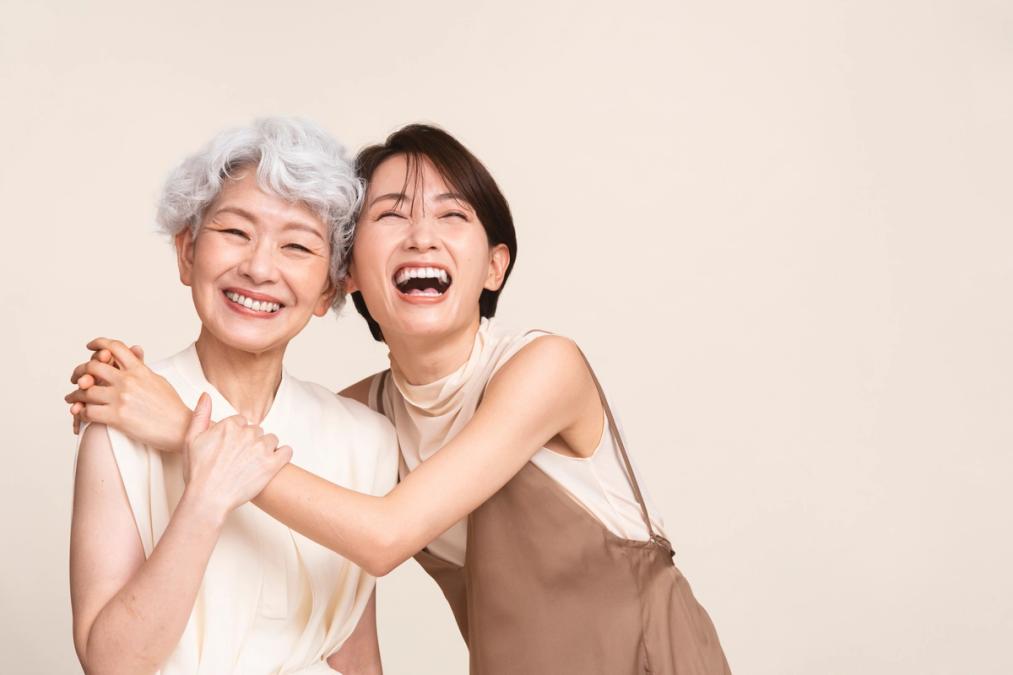 Two Asian women, one older with grey hair and one younger, embrace and laugh heartily.