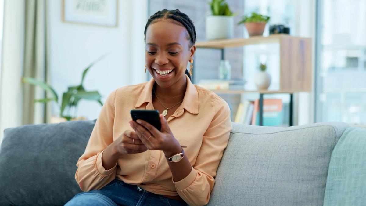 A smiling Black woman on a sofa using a smartphone.