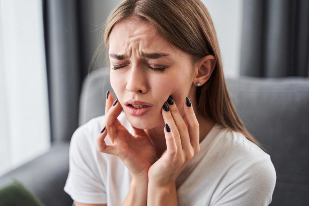 A young woman with her eyes closed, grimacing and holding her jaw due to pain.