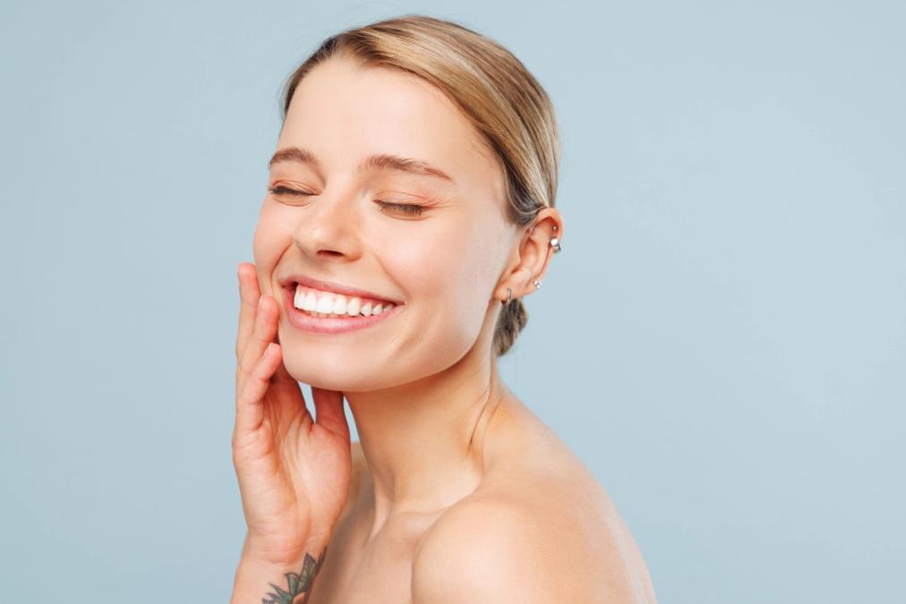 A smiling woman with closed eyes gently touches her cheek.