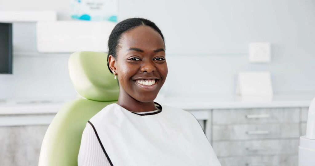A smiling Black woman sits in a dental chair wearing a protective bib.