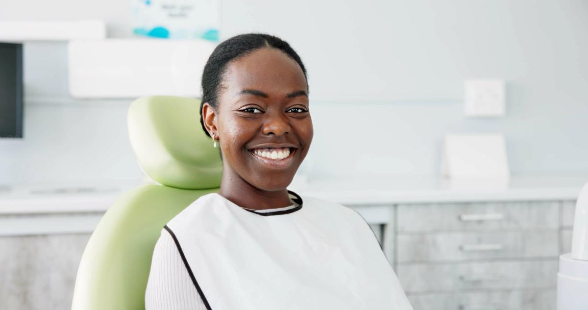 A smiling Black woman sits in a dental chair wearing a protective bib.