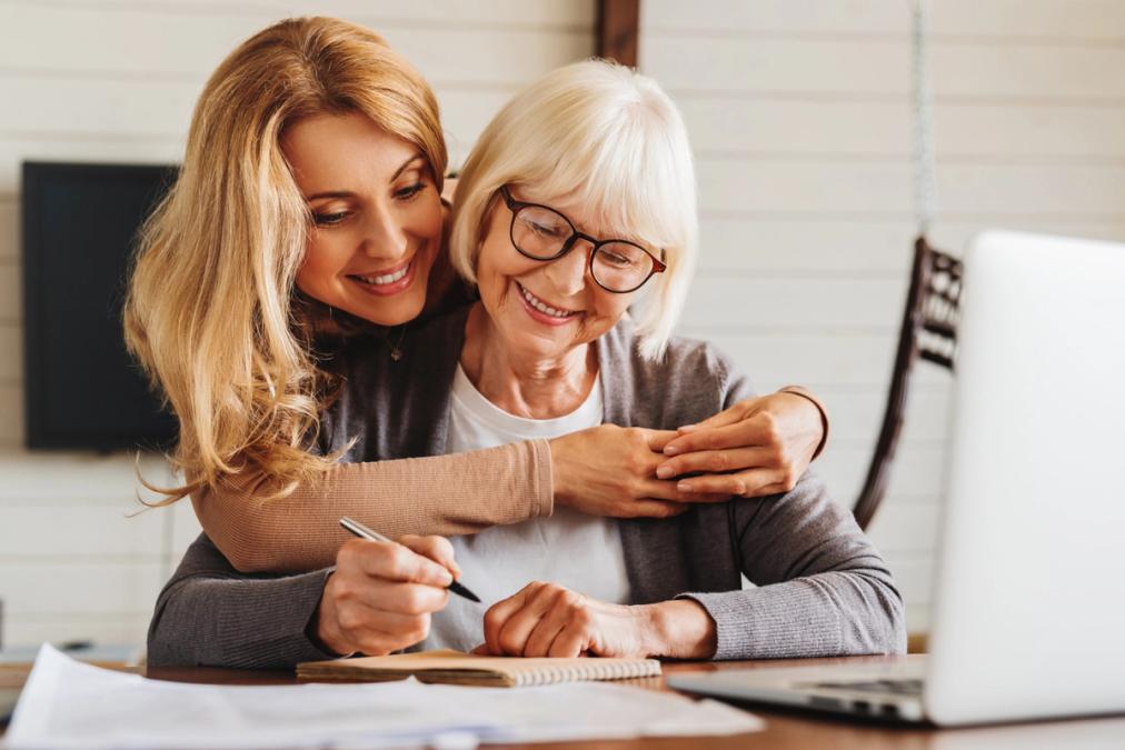 A smiling young woman embraces an older woman who is writing in a notebook.