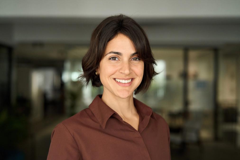 A smiling woman with short dark hair wears a brown shirt in an office setting.