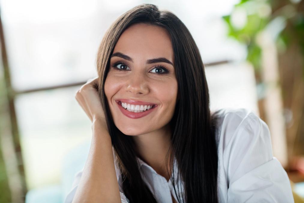 Smiling woman with long dark hair, wearing a white shirt, resting her hand on her chin.