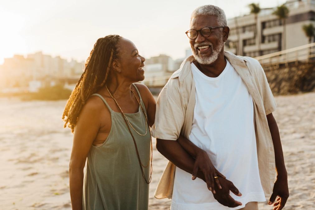 An older couple smiles while walking arm-in-arm on a sunny beach.
