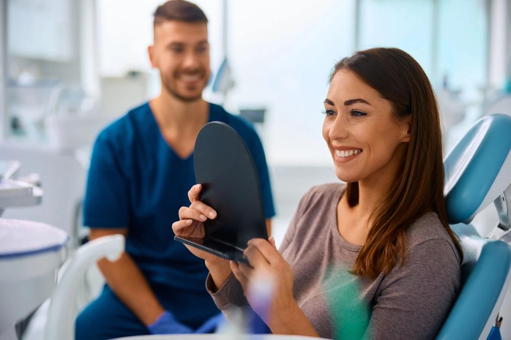 A happy woman in a dental chair smiles at her reflection in a mirror, while a smiling dentist watches in the background.